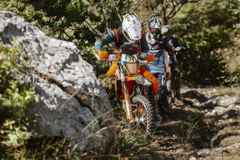 Riders pushing a dirt bike past a boulder in the forest